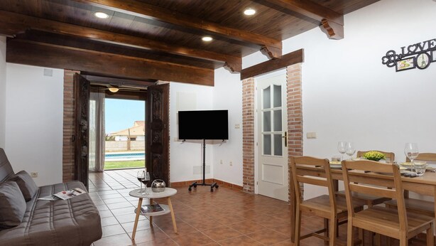 A living area with a sofa, a dining table set for four, a TV mounted on the wall, and a door leading to a balcony.