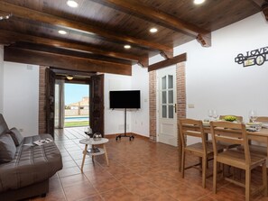 A living area with a sofa, a dining table set for four, a TV mounted on the wall, and a door leading to a balcony.