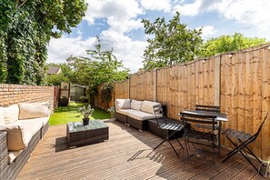 A backyard with a wooden deck, a sofa, a table, and a garden area. - Stylish Family Home with Garden in Peckham Rye (London)
