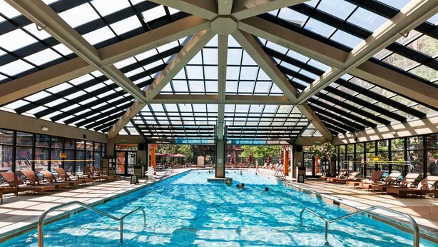 An indoor swimming pool with a glass ceiling, surrounded by lounge chairs and a tiled floor.