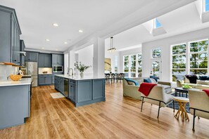 A spacious kitchen with grey cabinets and a large island, a dining area with a table and chairs, and a living room with a sofa and a coffee table.