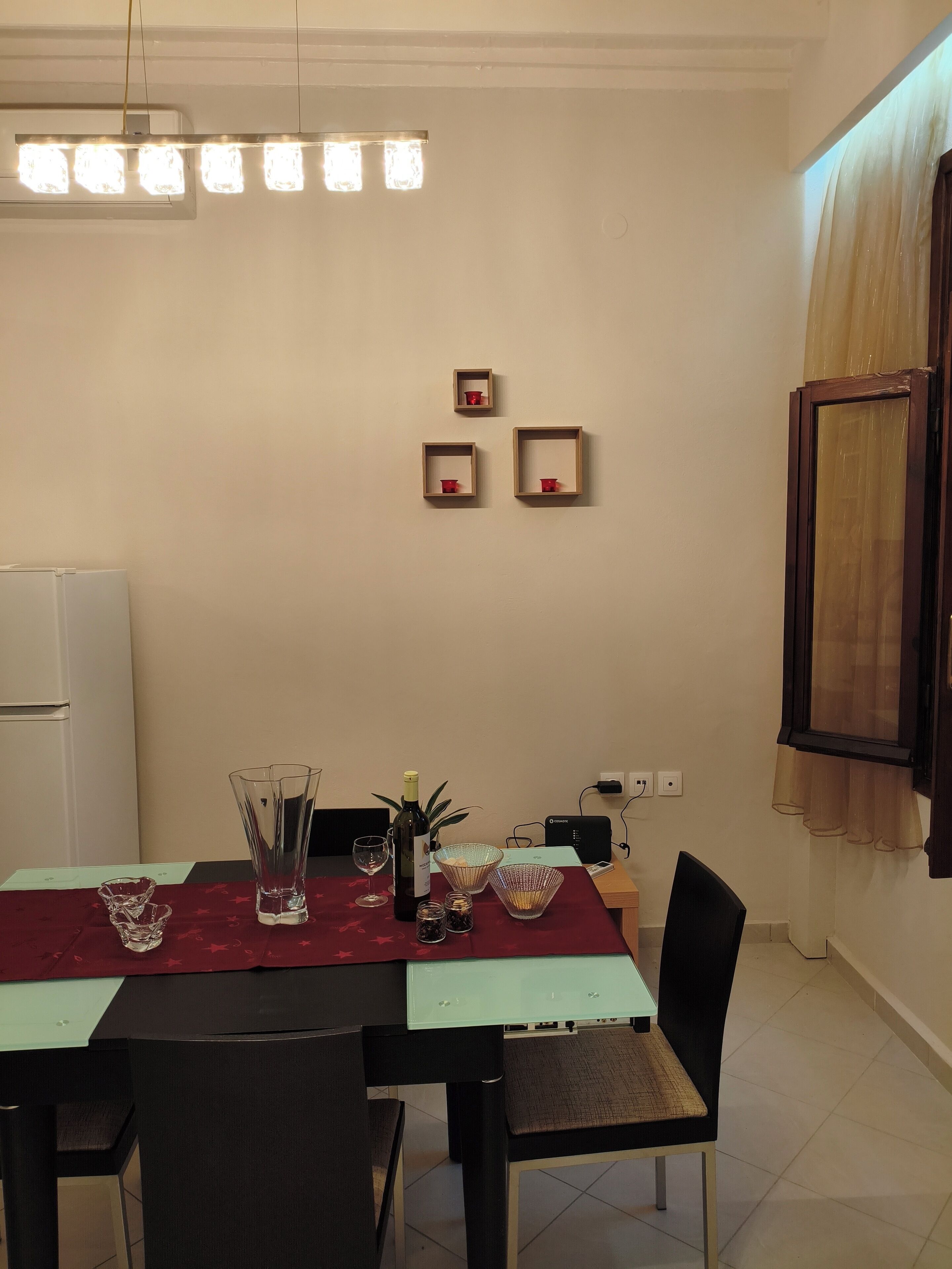 A dining area with a table set for four, a refrigerator, a plant, and a window with curtains.