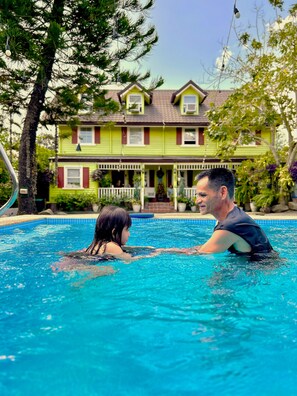 A swimming pool with two people in it. In the background, there is a house with a porch and a tree. - Madie's Place B&B with Private Pool in Santa Rosa near Enchanted Kingdom (City of Santa Rosa)