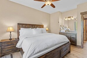 A bedroom with a wooden bed, two bedside tables with lamps, a ceiling fan, and a bathroom with a vanity and mirror.
