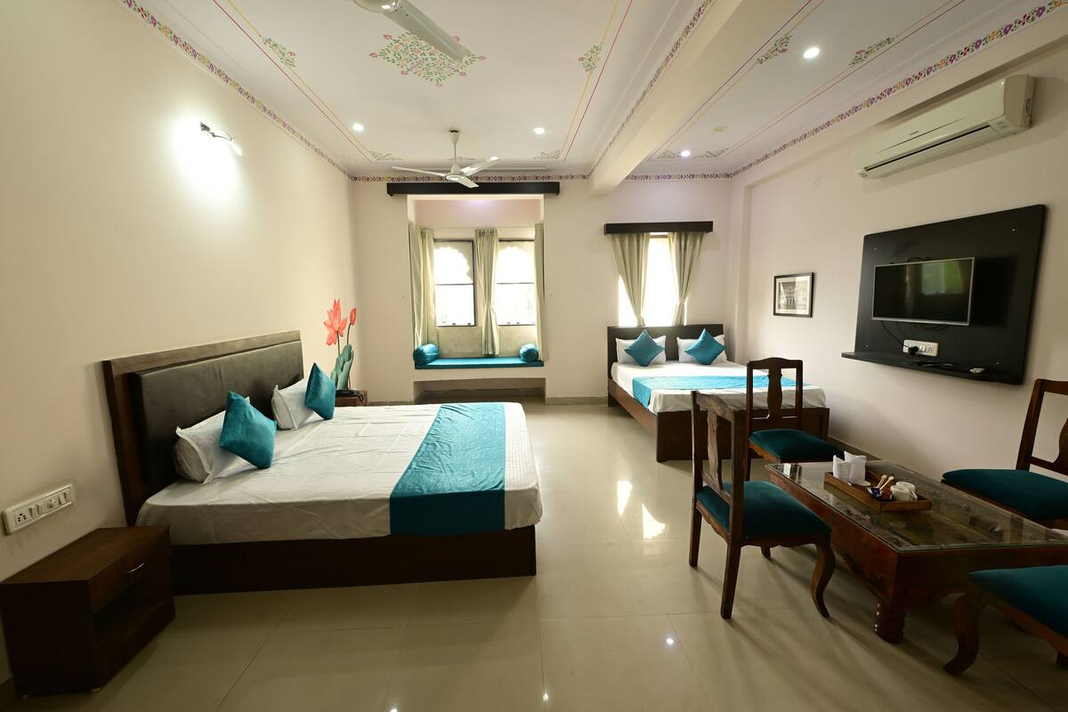 A hotel room with a large bed, a smaller bed, a desk with chairs, a television, and a ceiling fan.