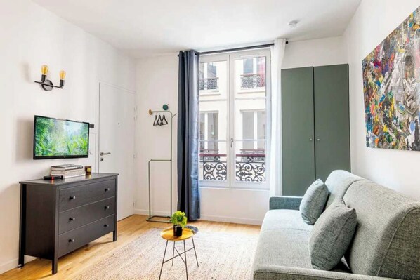 A modern living room with a grey sofa, a television, a small table with a plant, and a balcony with a view.