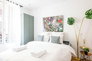 A bedroom with a bed, pillows, a painting, a plant, and a wardrobe.