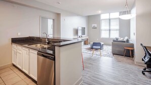 A modern kitchen with a dishwasher, a dining area with a sofa and a TV, and a large window providing natural light. - TWO CozySuites at the Banks 17 (Cincinnati)