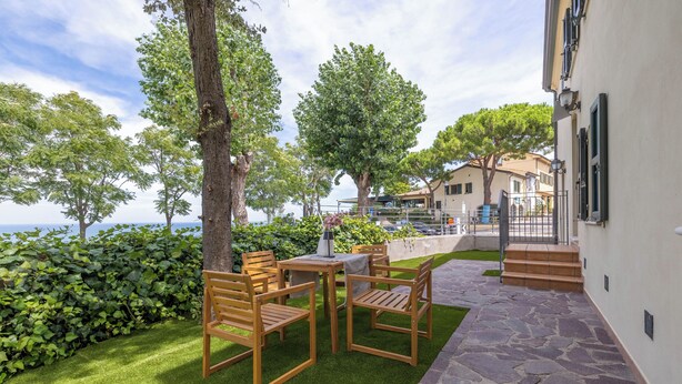 An outdoor patio with wooden chairs and a table, surrounded by greenery and a stone pathway. - La Finestra sul Mare - Apartment with a view (Gabicce Mare)