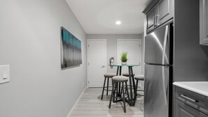 Une cuisine moderne avec un réfrigérateur en acier inoxydable, une table à plateau en verre et des tabourets, et une porte donnant sur une autre pièce.