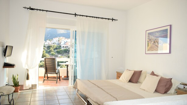 A bedroom with a bed, a chair, a TV, and a balcony with a view of buildings.