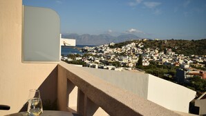 A balcony with a table, a glass of wine, and a view of a coastal town with buildings and mountains in the background.
