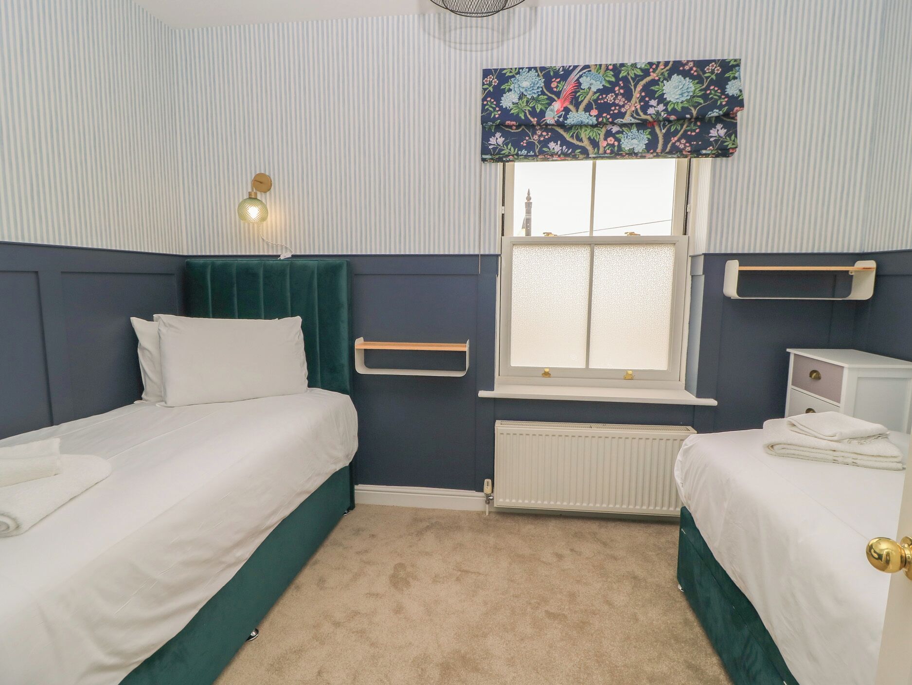 Habitación de hotel con dos camas, una ventana con vistas, un radiador y una mesita de noche con lámpara.
