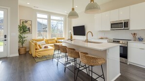 A modern kitchen with white cabinets, a large island with bar stools, a flat-screen TV, and a dining area with yellow furniture. - 2 New Townhomes w/ Balcony, Mins to Broadway, Sleeps 16 | 98 Degrees & The Stage (Nashville)