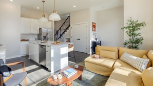 A modern kitchen with white cabinets, a central island, and pendant lighting. There is a yellow sofa with a plant beside it. - 2 New Townhomes w/ Balcony, Mins to Broadway, Sleeps 16 | 98 Degrees & The Stage (Nashville)