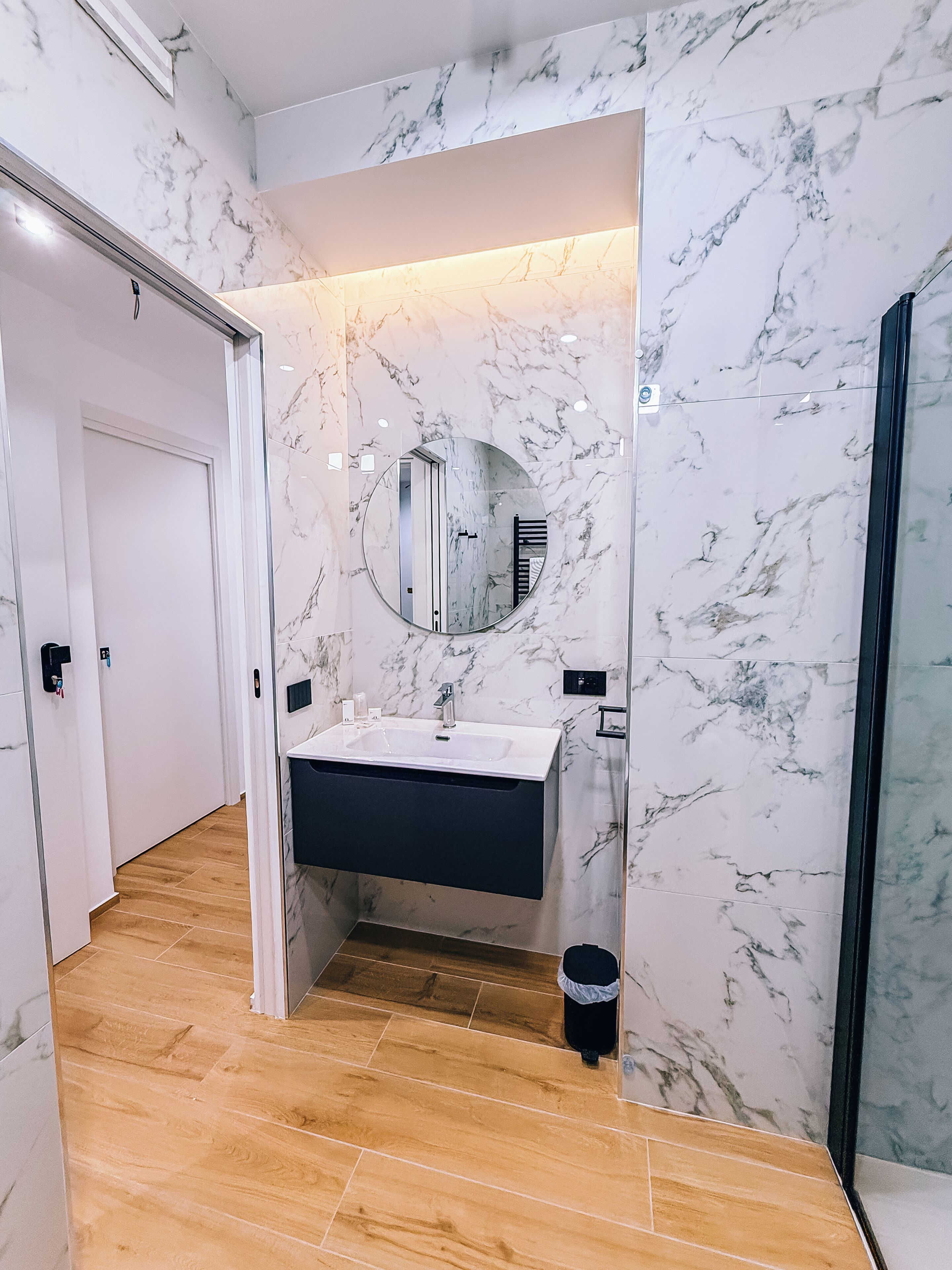 Un baño moderno con pared de mármol, un espejo redondo, un lavabo y zona de ducha.