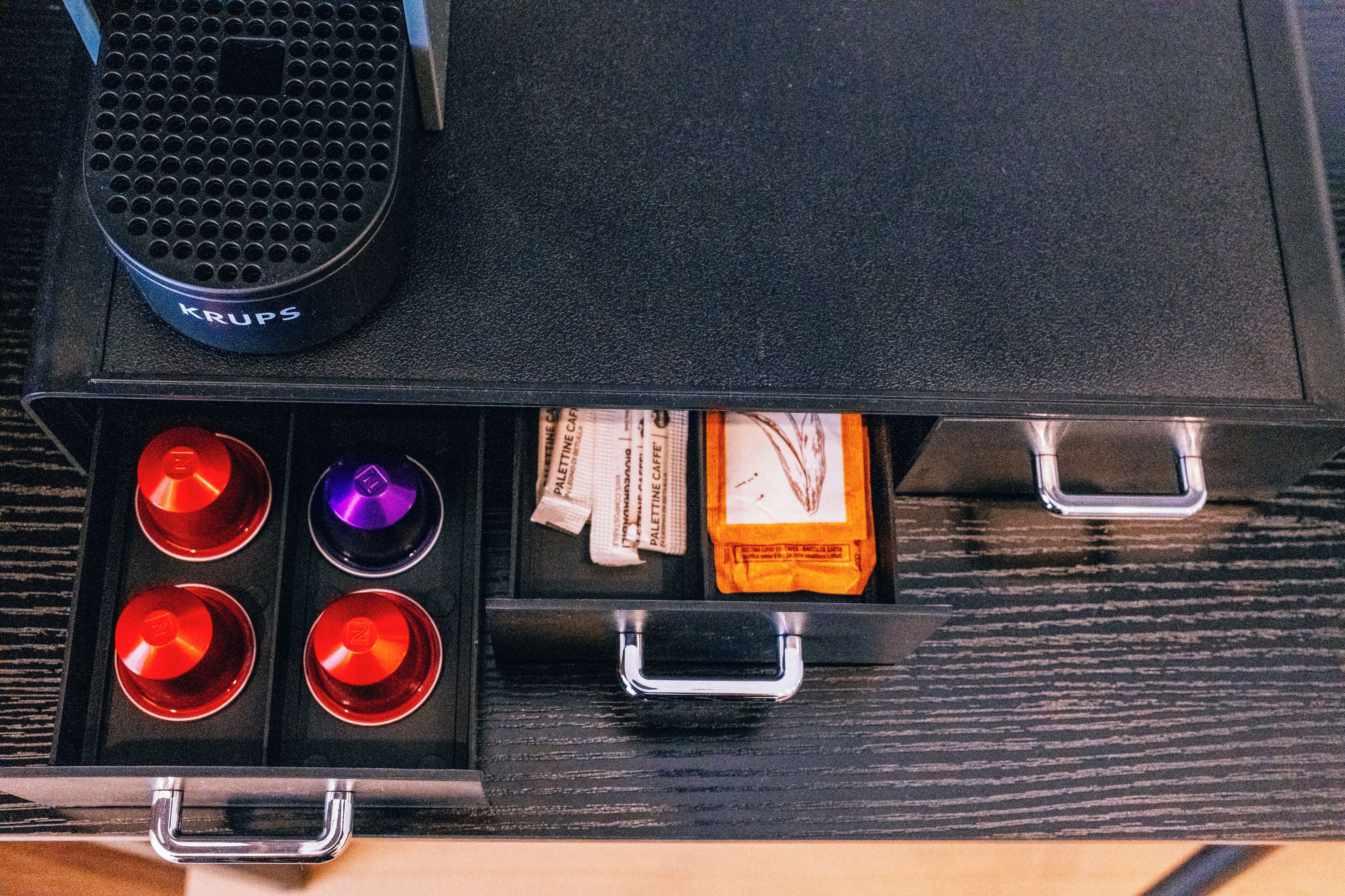 A drawer with compartments containing various items including a Krups coffee maker, red and purple lids, and packaged food items.