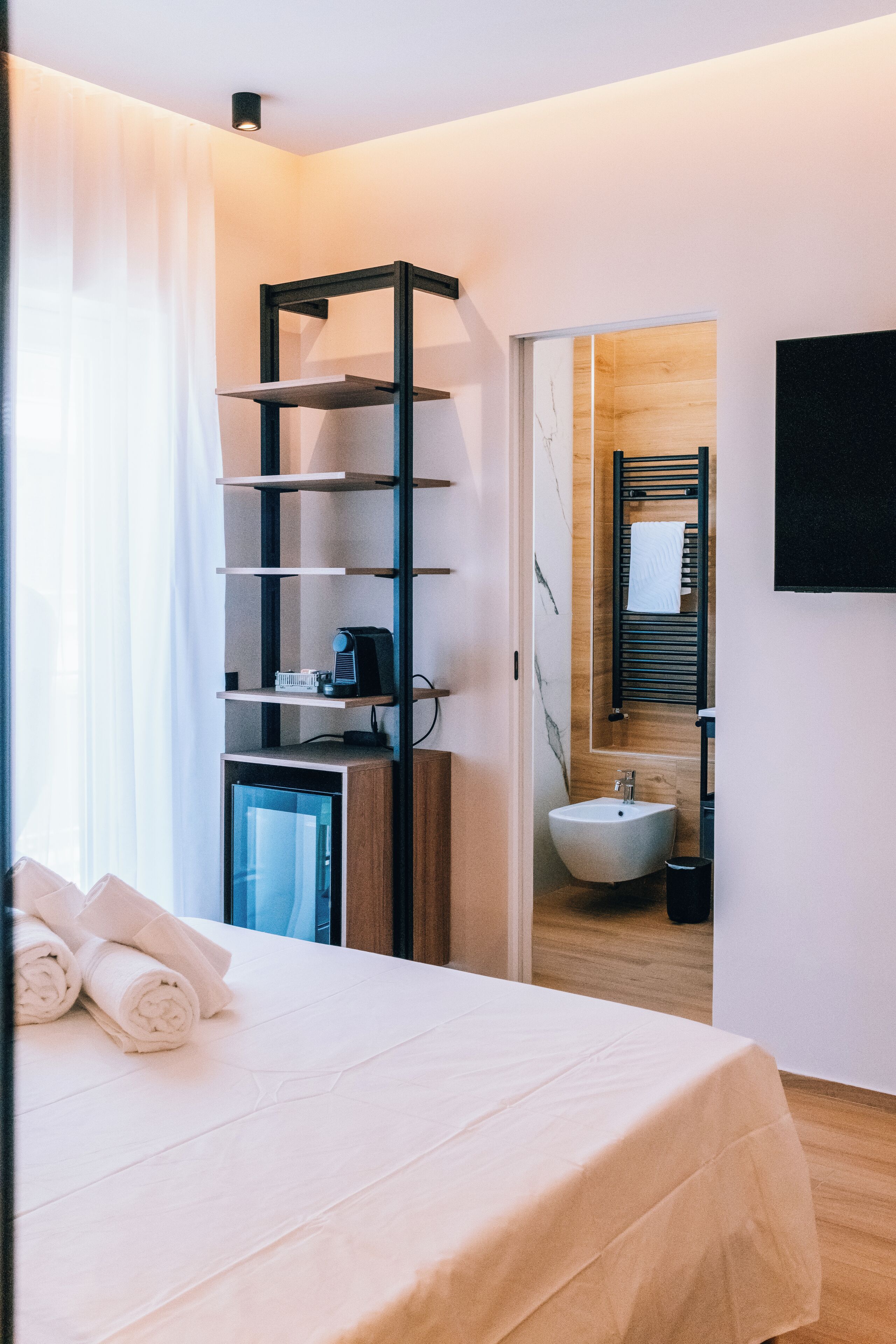 Une chambre d’hôtel comprenant un lit, une table de chevet avec une lampe, une étagère intégrée, une salle de bain avec un lavabo et un miroir, et un téléviseur fixé au mur.