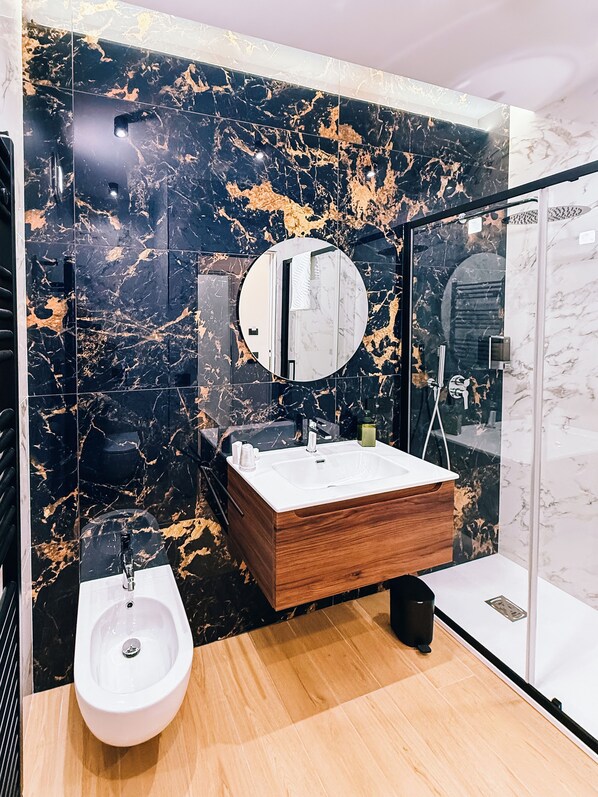 A modern bathroom with a marble backsplash, a wooden vanity, a white sink, a bidet, and a glass shower enclosure.