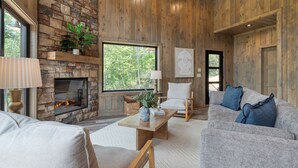 Ein gemütliches Wohnzimmer mit Kamin aus Stein, Holzwänden und einem großen Fenster mit Blick auf Bäume.