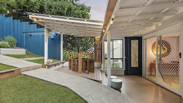A covered patio area with a wooden table and chairs, a concrete pathway, and a blue fence in the background.