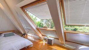 Un dormitorio con techo inclinado, suelo de madera, cama, mesita de noche y ventana con persianas.