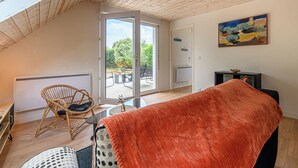 Una habitación con techo inclinado, un ventanal grande, una silla de mimbre, una mesa y una manta roja.