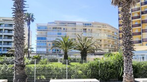 A modern multi-story building with balconies, surrounded by palm trees and a metal fence.