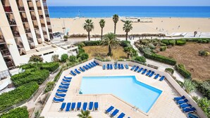 A pool area with blue lounge chairs, a clear view of the beach, and a building with balconies.
