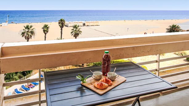 A balcony with a table set for breakfast, overlooking a beach with palm trees and the ocean.