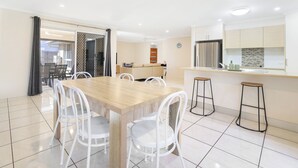 A modern dining area with a wooden table and white chairs, a kitchen with a stainless steel refrigerator, and a sliding glass door leading to an outdoor area. - Bargara Haven
Welcome to your personal oasis: Coastal calm blends with style. (Bargara)
