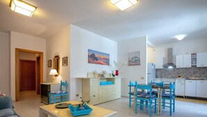 Un salon moderne avec un canapé, une table basse et un coin repas comprenant des chaises bleues et une table. On y trouve également une cuisine avec des meubles blancs et une cuisinière.