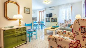 Un salon comprenant un meuble vitré, un canapé recouvert d’un tissu à motifs floraux, un coin repas avec des chaises bleues et un téléviseur posé sur un meuble.