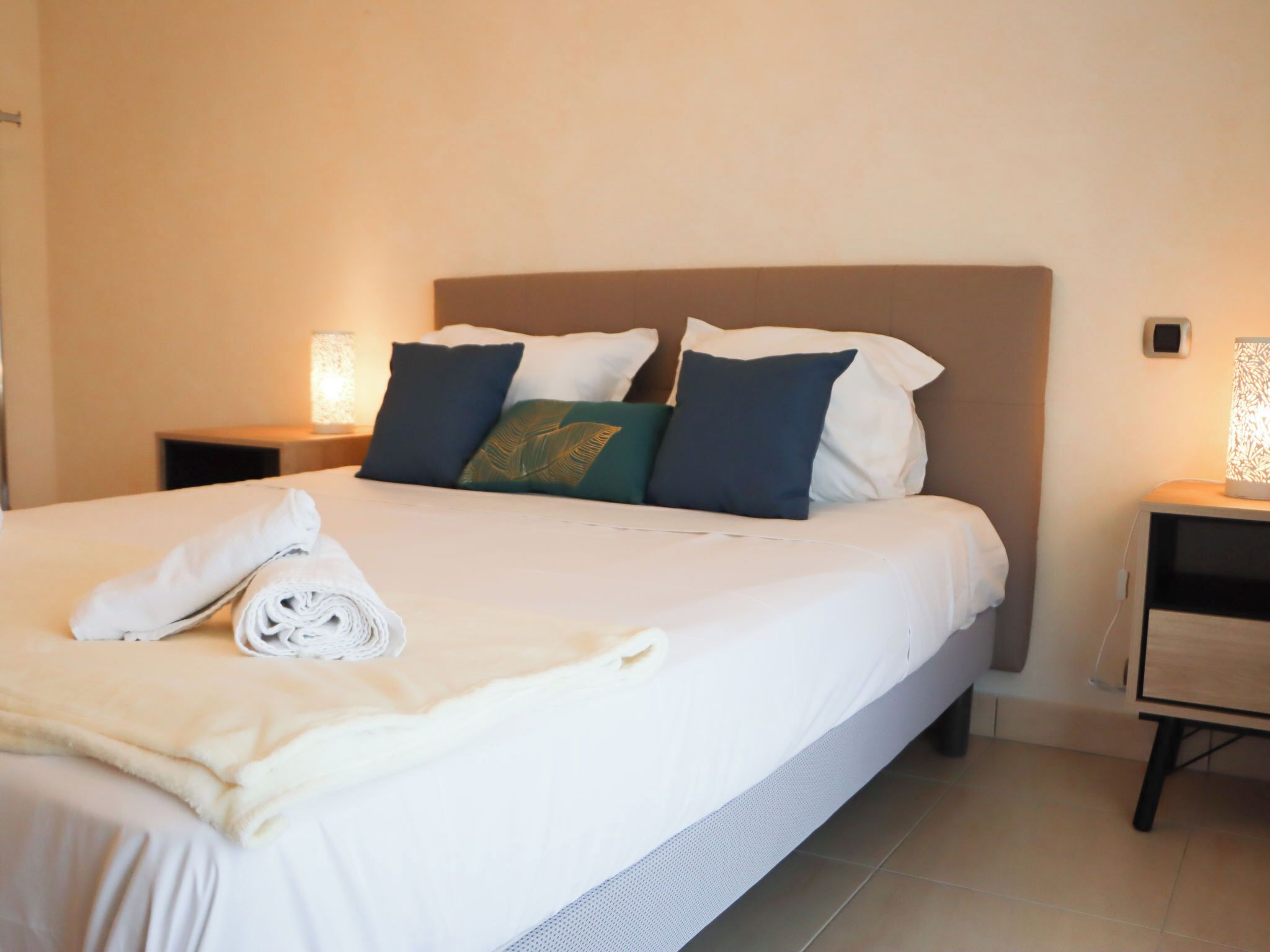 A neatly made bed with white linens, a folded towel, and a decorative pillow.