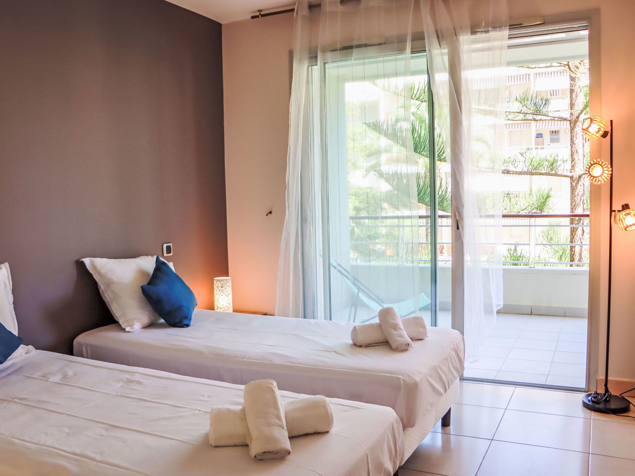 A hotel room with two beds, a balcony with a view of greenery, and a floor lamp.