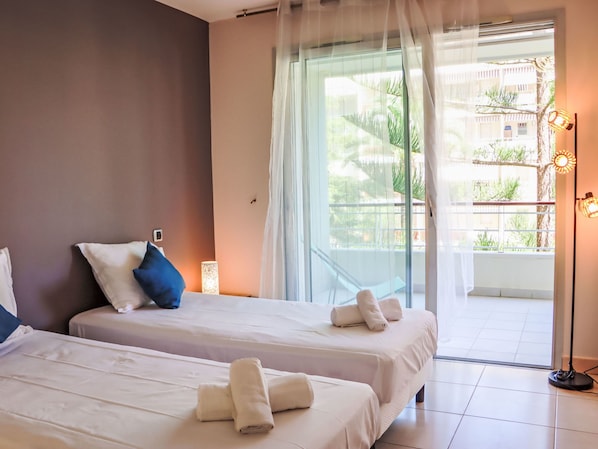 A hotel room with two beds, a balcony with a view of greenery, and a floor lamp.