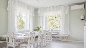 Uma área de refeições com mobiliário branco, grandes janelas com cortinas leves e vista para o verde lá fora.