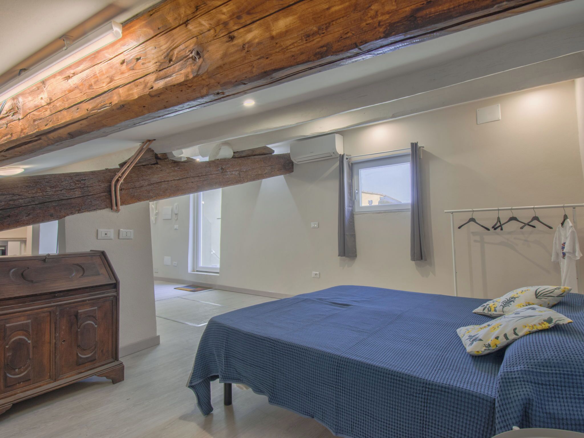 Une chambre avec un plafond en bois, un lit recouvert d’une couverture à carreaux bleus, une commode en bois et une fenêtre avec des rideaux.