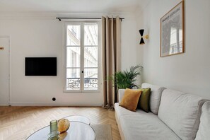 Uma sala de estar com sofá, televisão, janela com cortinas, uma planta e um quadro na parede.