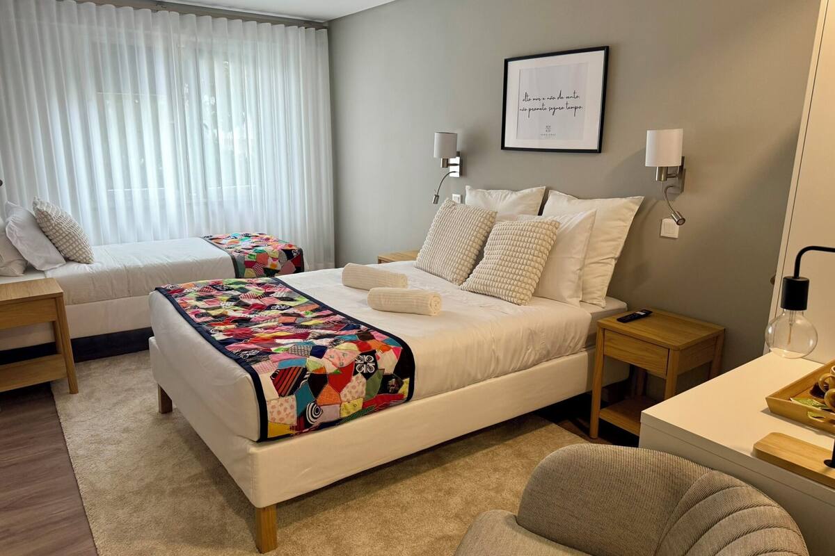 Une chambre d’hôtel avec un grand lit, deux tables de chevet, un bureau, une chaise, une fenêtre avec des rideaux, une lampe fixée au mur et une citation encadrée accrochée au mur.