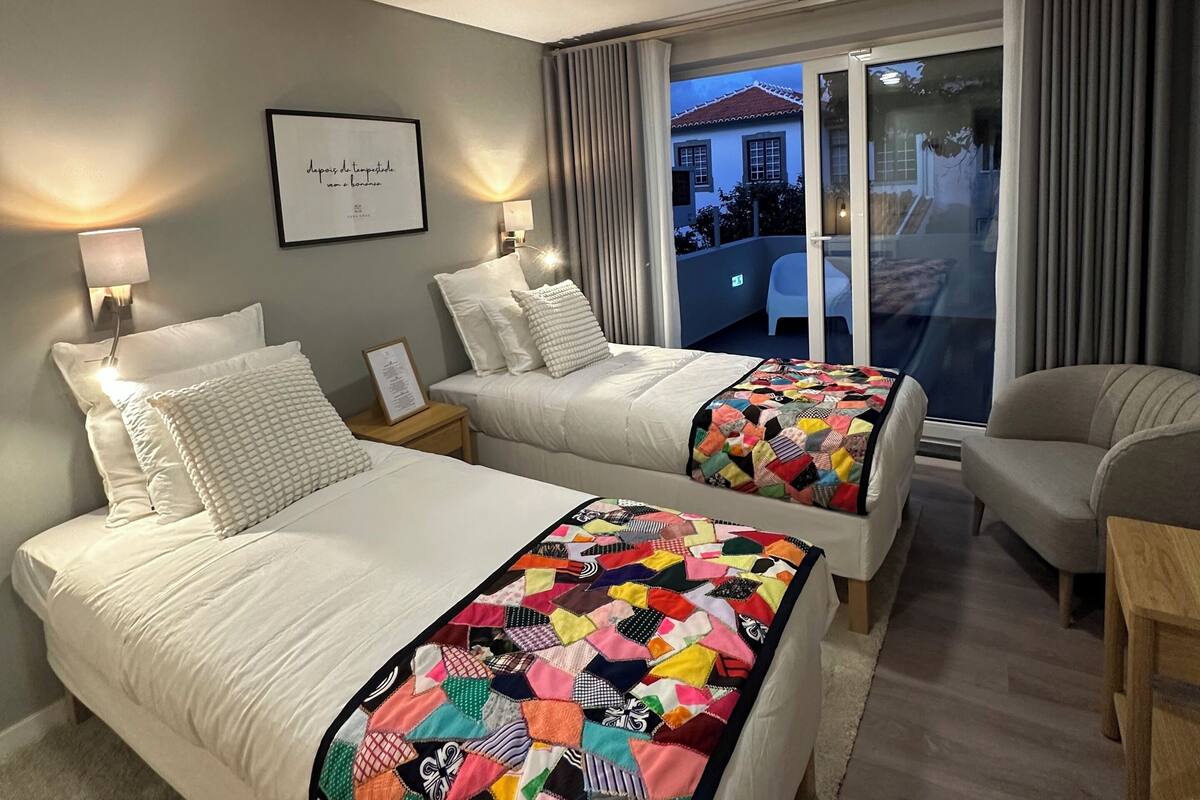 Une chambre d’hôtel avec deux lits, un canapé, un fauteuil et une vue sur un immeuble, visible à travers une porte vitrée.