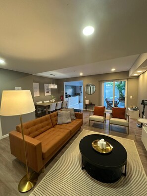 A modern living room with a sofa, coffee table, and a lamp. There is a kitchen in the background with a dining area.