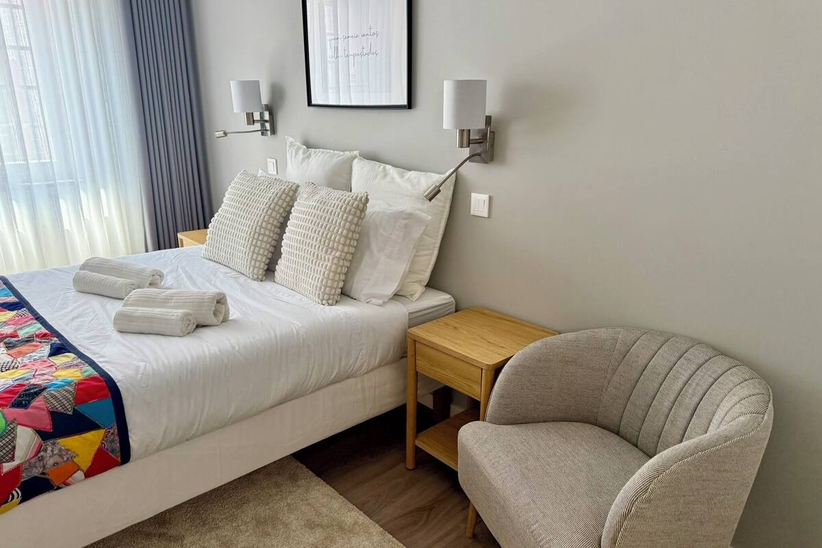 Un lit bien fait, avec du linge de lit blanc et une couverture colorée, un fauteuil gris, une table de chevet en bois et deux lampes fixées au mur.
