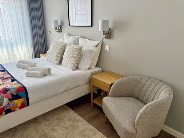 A neatly made bed with white bedding and a colorful quilt, a gray armchair, a wooden nightstand, and two wall-mounted lamps.