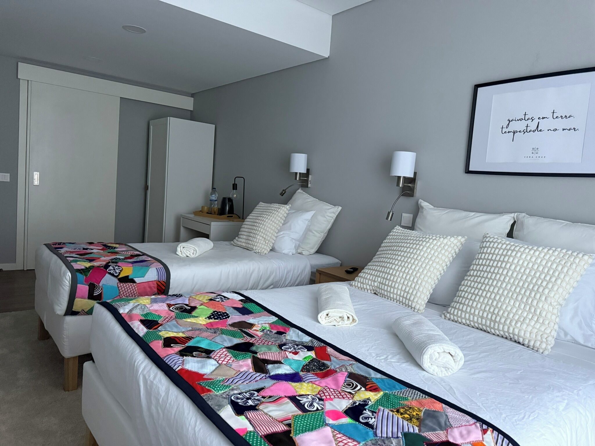 Habitación de hotel con dos camas, colcha colorida, ropa de cama blanca, una cita enmarcada en la pared y una mesita de noche pequeña con lámpara.