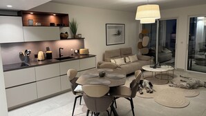 Un espace de vie moderne comprenant une cuisine, un coin repas et un coin salon. Il est équipé d’un canapé, de fauteuils, d’une table à manger et d’un comptoir de cuisine avec des appareils électroménagers.