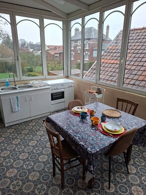 A sunroom with a dining table set for two, a kitchen area with a sink and stove, and large windows offering a view of the neighborhood.