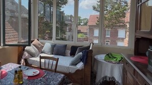 A cozy room with a sofa, a dining table, and a view of a brick building through large windows. - 1 studio apartment for rent in Villa Amenaide: 1 bedroom, plus kitchen, dining room, and living room (Roisel)