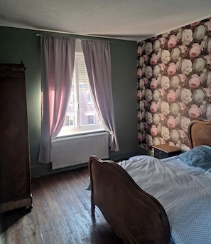 A bedroom with a wooden bed, a radiator, a window with curtains, and a floral wallpaper.