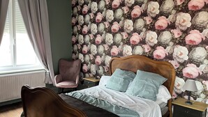 A bedroom with a floral wallpaper, a wooden bed, a chair, a bedside table with a lamp, and a window with curtains. - 1 studio apartment for rent in Villa Amenaide: 1 bedroom, plus kitchen, dining room, and living room (Roisel)